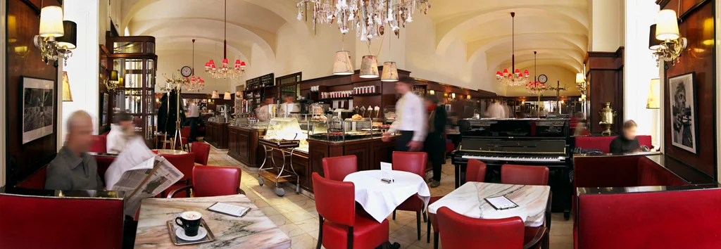 Der weite Blick durch das Café Diglas: rote Polsterbänke, Marmortische, Klavier und Kaffeehausgäste
